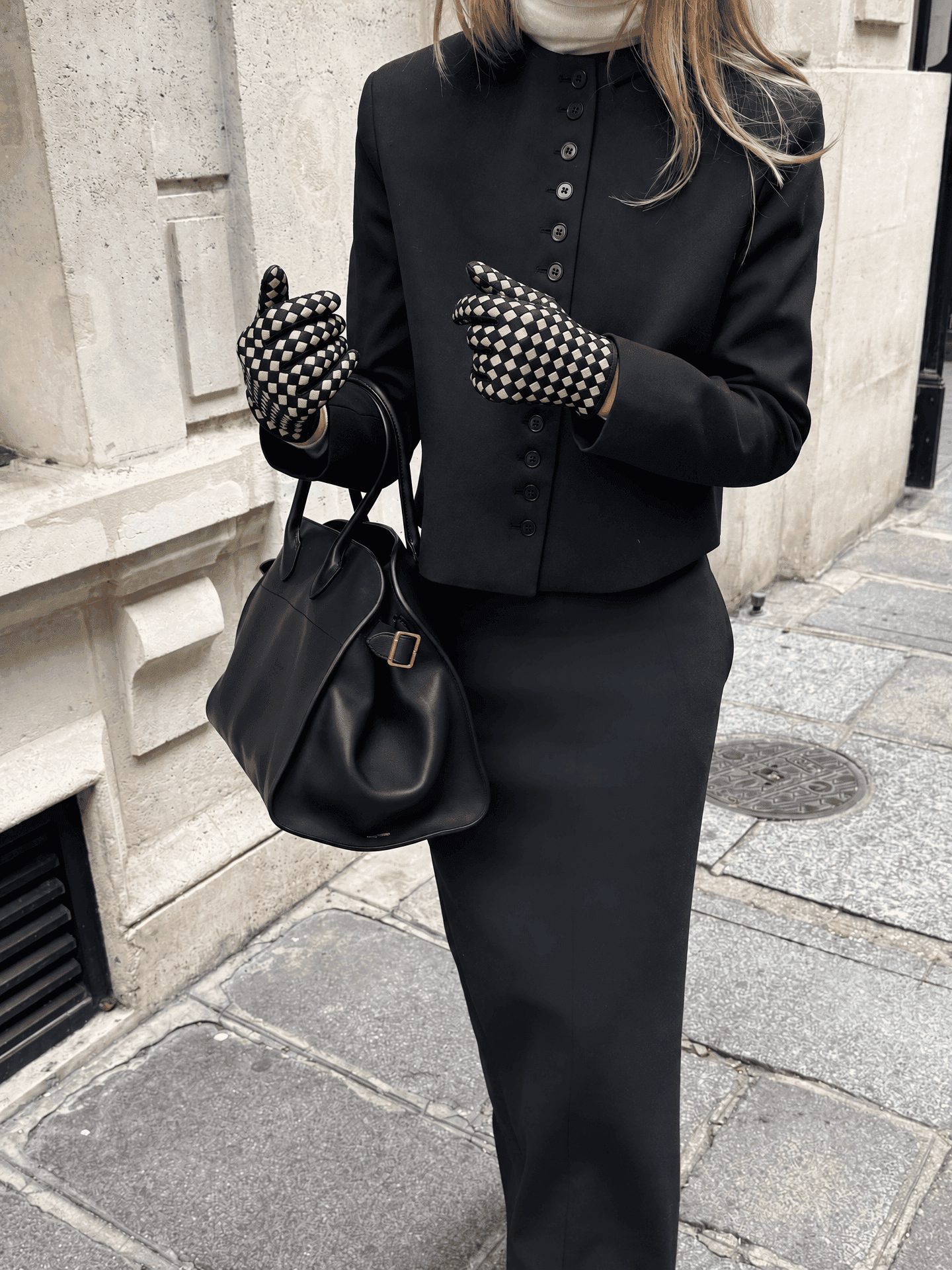 a woman on a paris pavement in a black buttoned jacket, a cream roll-neck and a long black skirt, carrying a black leather bag and wearing black suede gloves with a cream checkerboard
