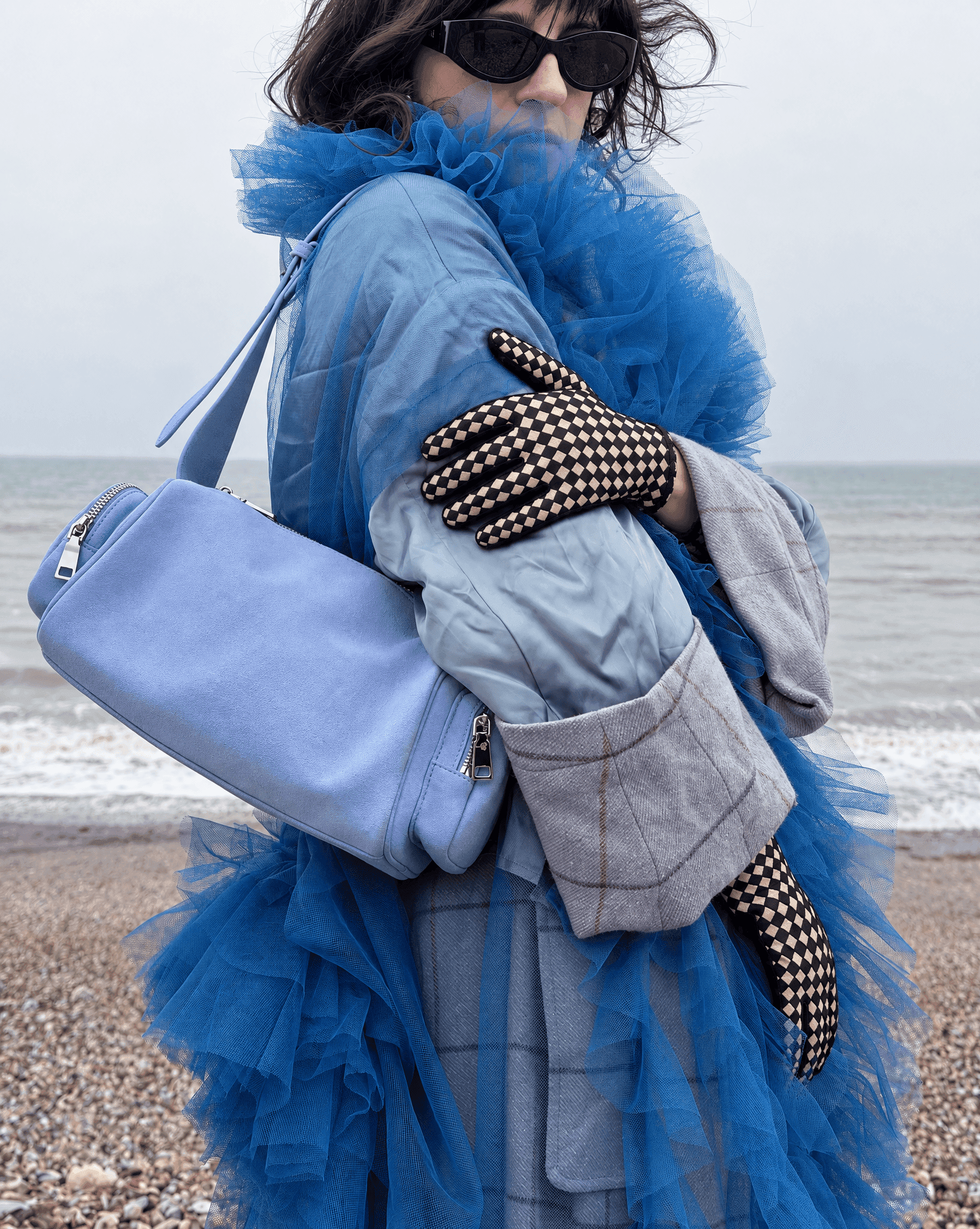 black and cream checkerboard suede gloves on a woman in a pale blue coat and blue tulle, holding a periwinkle bag on a pebble beach