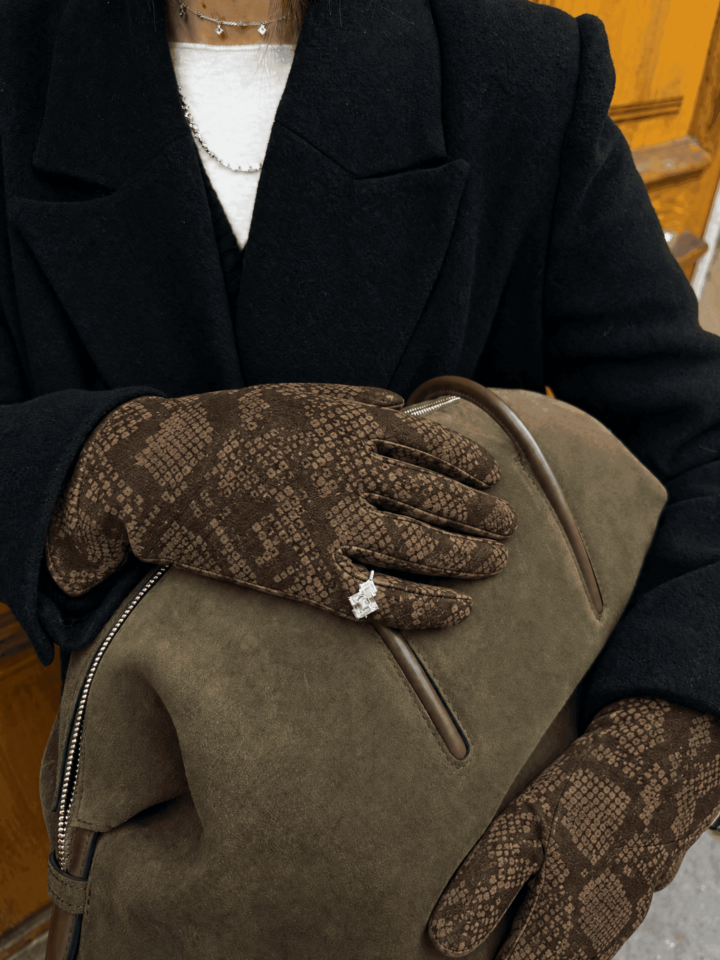 a silver ring worn over brown suede gloves printed with a python pattern, with a black coat and an olive suede bag